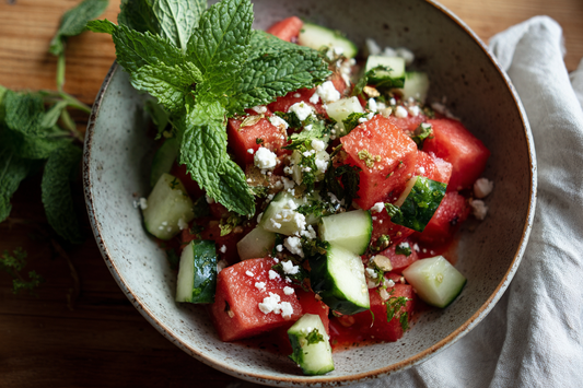Sommerlicher Melonen-Gurken-Salat mit frischer Minze, Limette und veganem Feta in einer grauen Schüssel auf einem Holztisch, perfekt für warme Tage.