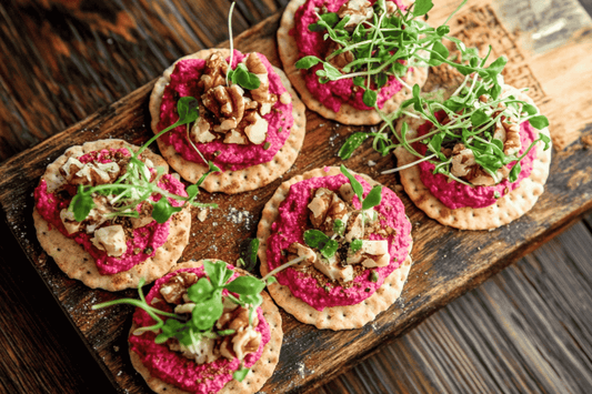 Kleine Cracker mit leuchtend pinkem Rote-Bete-Hummus, dekoriert mit Walnüssen und frischer Kresse, auf einem rustikalen Holzbrett angerichtet
